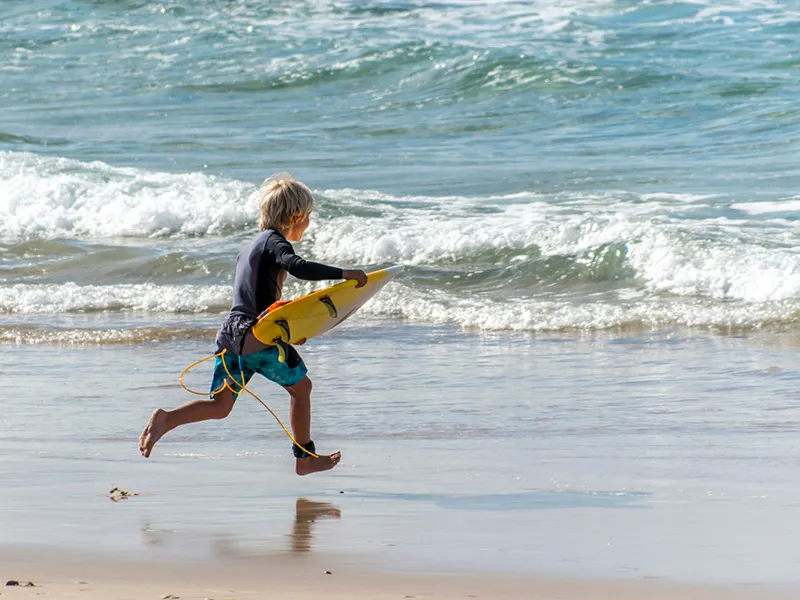 Enfant courant sur la plage avec une planche de surf jaune, initiation surf en famille.